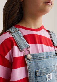 Laps, kes kannab punaseid ja roosasid triibulist särki ning hele sinist teksast totaaliga, millel on eesmine tasku ja metallist pandlaga rihmad.