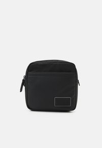 Borsa a tracolla nera in nylon con forma rettangolare, dotata di una tasca frontale con zip e un'etichetta con logo. Texture liscia, design minimalista.
