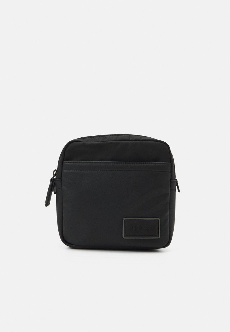 Borsa a tracolla nera in nylon con forma rettangolare, dotata di una tasca frontale con zip e un'etichetta con logo. Texture liscia, design minimalista.