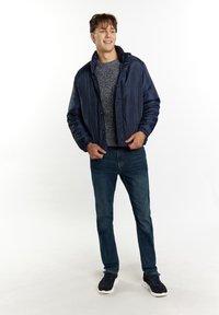 Giubbotto imbottito blu navy con colletto alto e bottoni a pressione, indossato sopra un maglione grigio a trama, abbinato a jeans blu e sneakers nere.