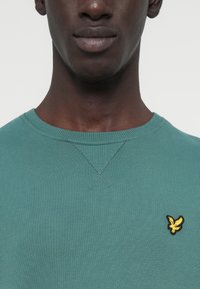 Τιρκουάζ φούτερ με στρογγυλή λαιμόκοψη και ραβδωτή υφή, με μικρό κίτρινο κεντητό λογότυπο στο αριστερό στήθος και ενισχυμένες ραφές στους ώμους.