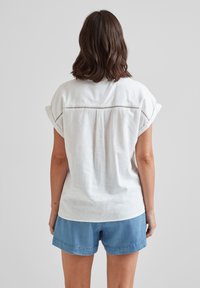White linen top with rolled sleeves and horizontal eyelet detailing across the upper back. Paired with blue denim shorts.