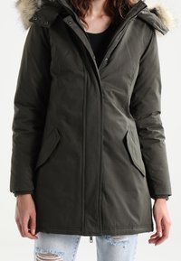 Parka vert foncé avec une capuche bordée de fourrure, fermeture éclair, poches latérales et poignets côtelés. Le matériau semble épais et résistant aux intempéries.