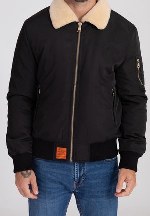 Mann trägt eine schwarze Bomberjacke mit cremefarbenem Lammfellkragen, Reißverschlusstaschen und blauen Jeans, steht vor einfachem Hintergrund.