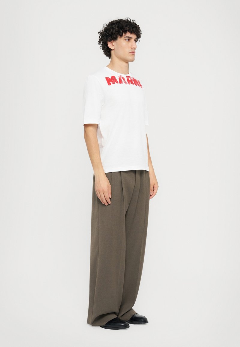 T-shirt blanc avec le texte rouge "MAFIA", associé à un pantalon taille haute, large, de couleur olive, et des chaussures noires. Tissu finement tissé.