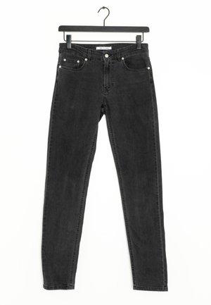 Czarne, wysokowciśnięte obcisłe dżinsy z przednimi kieszeniami, guzikiem i szlufkami na pasek, zawieszone na czarnym wieszaku na białym tle.