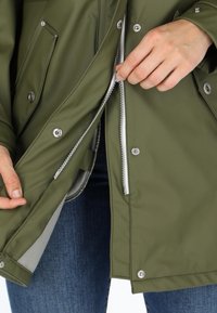 Giacca impermeabile verde oliva con cerniera argento e bottoni a pressione, con una texture liscia e un interno foderato, parzialmente zipata.
