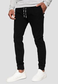 Man som bär svarta joggarbyxor med dragsko och vita sneakers, står med händerna i fickorna mot en grå bakgrund.