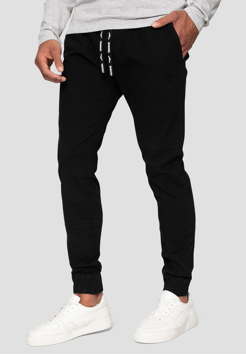 Man som bär svarta joggarbyxor med dragsko och vita sneakers, står med händerna i fickorna mot en grå bakgrund.