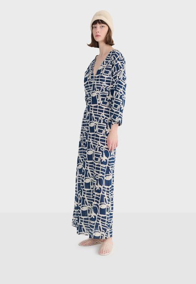 Kobieta ubrana w długą granatową sukienkę z białymi abstrakcyjnymi wzorami, beżową dzianinową czapkę i jasne, plecione buty wsuwane, stojąca bokiem.