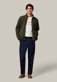 Oliivinvihreä fleecetakki, jossa on kaksi suurta etutaskua, valkoinen t-paita, jossa on vuoristografiikka, tummansiniset housut ja ruskeat mokkakengät.