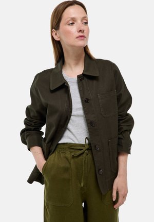 Femme portant une veste boutonnée vert foncé sur une chemise gris clair et un pantalon vert assorti, les mains dans les poches, regardant sur le côté.