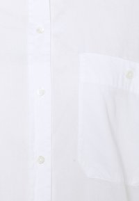 Chemise blanche à boutons, confectionnée en tissu léger, avec une petite poche sur la poitrine gauche et des boutons en plastique transparent. Texture lisse.