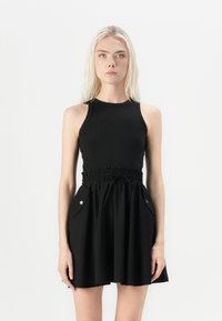 Svart kjole med tettsittende ribbestrikket overdel, smocket midje, og vide skjørt med sidelommer og trykknapper. Jevn stoffoverflate.