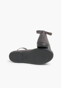 Zapato de ante gris con forma curvada y correa en el tobillo que presenta una suela de goma negra texturizada. Acento de hebilla de metal en la correa.