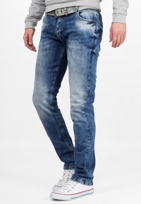 Blauwe denim jeans met een slim fit, met lichte slijtage, vijf zakken en een grijze riem. Gecombineerd met witte sneakers.