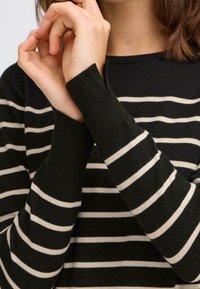 Maglione a righe nere e crema con texture a coste, caratterizzato da maniche lunghe e una scollatura aderente, con particolare attenzione alla posizione delle mani vicino al viso.