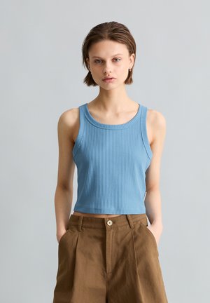 Młoda osoba z krótkimi włosami nosi niebieski, prążkowany, bezrękawnikowy top typu crop oraz brązowe spodnie z wysokim stanem, ręce w kieszeniach, neutralne tło.