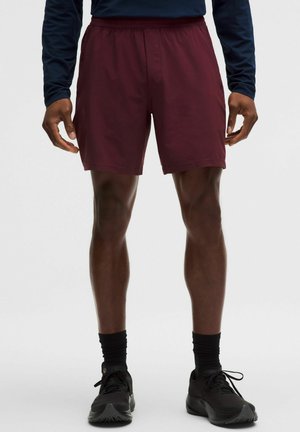 Mężczyzna ubrany w bordowe krótkie spodenki sportowe, czarne skarpetki i czarne buty do biegania, stojący na tle jednolitego jasnoszarego tła.