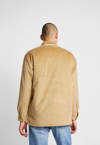 Brun kordjacka med klassisk krage, ribbad textur och knäppte ärmar. Bärs över ljusblå jeans och visar en avslappnad passform.