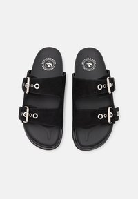 Sandali in suede nero con due cinturini regolabili, dotati di hardware argentato e ritagli circolari su una suola nera texturizzata. Logo del brand visibile.