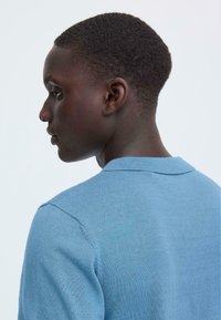 Polo bleu clair, tissu tricoté, manches courtes, col côtelé, texture lisse, coutures latérales visibles. Modèle présenté de dos.