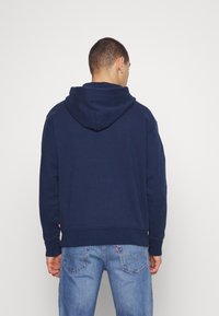 Tmavě modrá mikina s kapucí na šňůrku, žebrovanými manžetami a lemem. Měkká tkanina s kapsou na spodním lemu a uvolněným střihem.