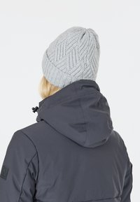 Berretto in maglia grigia con un motivo a spina di pesce testurizzato, indossato con una giacca scura dotata di cappuccio e di un dettaglio in patch sulla manica.