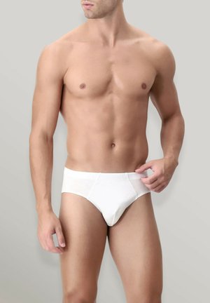 Uomo muscoloso con mutande bianche semplici, in piedi davanti a uno sfondo neutro, che sistema la cintura con la mano destra.