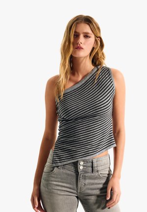 Ženska z dolgimi blond lasmi, oblečena v brezrokavnik s sivimi in črnimi diagonalnimi črtami ter svetlo sive jeans hlače, stoji pred enobarvno belo ozadje.