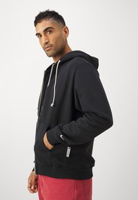 Sudadera negra con cremallera y cordón, bolsillo delantero, puños acanalados y un pequeño logo de Nike en la manga. Modelo que lleva pantalones rojos.