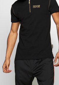 Zwarte polo met korte mouwen en een ritskraag, voorzien van gouden borduring "VERSACE JEANS COUTURE" en zichtbare stiksels.