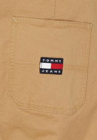 Tela beige con una textura suave que presenta un parche rectangular con "TOMMY JEANS" en blanco y rojo, cosido en un bolsillo.