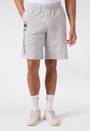 Mann trägt hellgraue Champion-Shorts mit Logo an der Seite, weiße Sportsocken und beige Sneaker, steht mit Händen in der Nähe der Taschen.