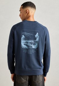 Mann med kort hår som har på seg en marineblå hettegenser med blå landskapsillustrasjon og tekst om å bevare naturlig arv på baksiden.