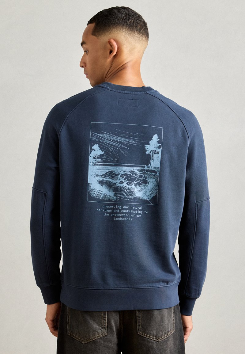 Mann med kort hår som har på seg en marineblå hettegenser med blå landskapsillustrasjon og tekst om å bevare naturlig arv på baksiden.