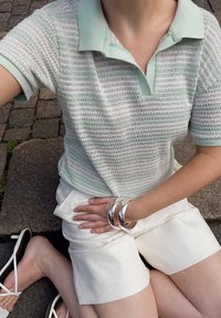 Polo majica s kratkimi rokavi v svetlo zeleni in beli črtasti vzorec z ovratnikom, v kombinaciji z belimi kratkimi hlačami in srebrnimi zapestnicami. Priložnostna obleka v sedečem položaju.