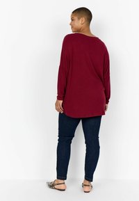 Bordó hosszú ujjú felső, lekerekített aljjal és laza szabással, sötétkék skinny farmernadrággal és ezüst kiegészítős szandállal párosítva.