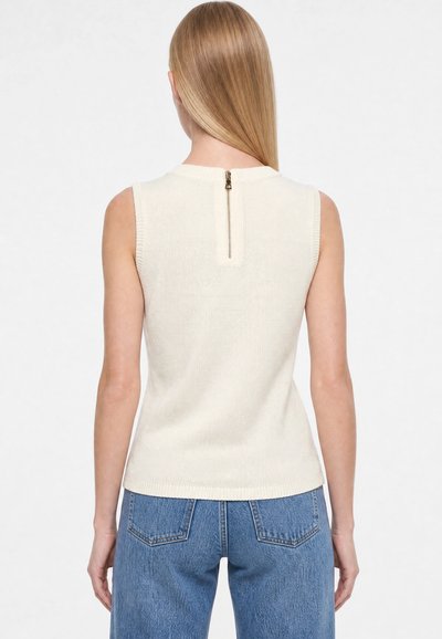 Kvinde med langt, lyst, glat hår iført en ærmeløs cremefarvet striktop med lynlås bagpå og blå denimjeans, set bagfra.