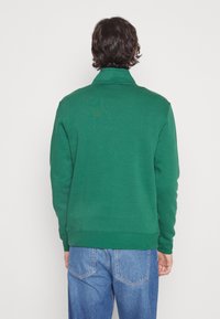 Zelená mikina s vysokým límcem, žebrovanými manžetami a lemem. Měkká látka s jednoduchou texturou. Nošeno s modrými džínami.