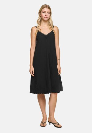 Woman with long blonde hair wears sleeveless black knee-length dress with scalloped edges and black sandals, standing against plain background.