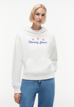 Ung kvinde med kort blondt hår iført hvid hættetrøje med teksten "Tommy Jeans" og røde stjerner, kombineret med blå jeans, stående mod en hvid baggrund.