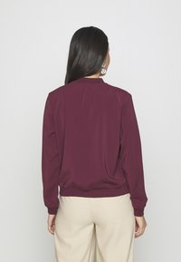 Burgundská bunda s dlouhými rukávy, volným střihem a žebrovaným lemem, vyrobená z hladké tkaniny. Zadní strana má jednoduchý design bez ozdob.