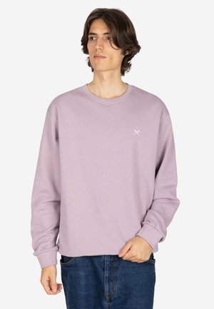 Junger Mann mit mittellangen braunen Haaren, der ein hellviolettes Sweatshirt und blaue Jeans trägt, steht vor einem schlichten weißen Hintergrund.