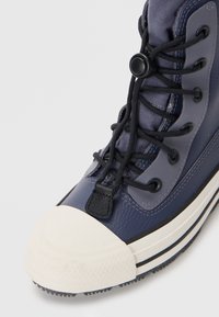Zapatilla alta azul marino con una puntera de goma blanca, cuero texturizado, cordones negros y un ojal negro redondo para un ajuste ajustable.