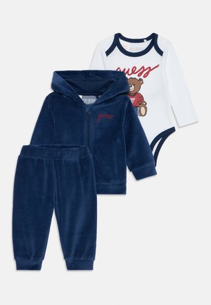 Tuta in pile blu navy con felpa con zip e pantaloni; body bianco presenta una grafica di un orso e il testo rosso "guess" lungo il girocollo.