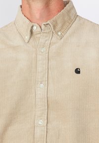 Camisa de pana beige con cuello abotonado, que presenta sutiles rayas verticales, botones blancos y un pequeño logo negro en el pecho.