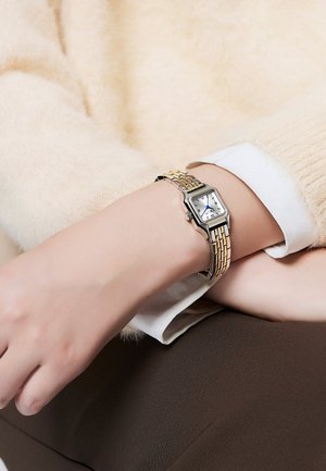Montre argentée et dorée au poignet. Cadran rectangulaire avec fond blanc, aiguilles bleues et bracelet en métal texturé. Un pull doux en arrière-plan.