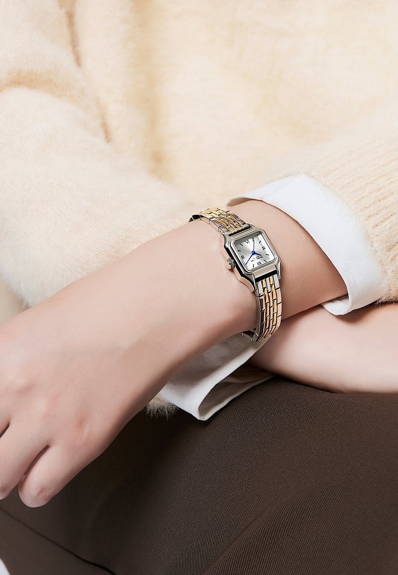 Montre argentée et dorée au poignet. Cadran rectangulaire avec fond blanc, aiguilles bleues et bracelet en métal texturé. Un pull doux en arrière-plan.