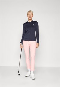Mornarsko modra zapenjala jakna z teksturirano tkanino, bela ovratnik, v kombinaciji s svetlo rožnatimi hlačami z vzorcem. Model drži golf palico, nosi bele športne copate.